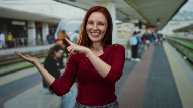 Tren istasyonunda gülümseyen, kırmızı giyen, elini gösteren, ayakta duran, dışarıda duran, kızıl saçlı, güzel, yetişkin, çekici, gösterişli seyahat ya da ulaşım sahnesi olan bir kadın..