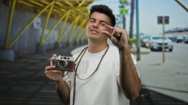 Latin asıllı genç bir adam elinde kamera ile şehir sokaklarında küçük bir jest yapıyor. Arabaları ve şehir arka planında, günlük beyaz gömlekler giyiyor..