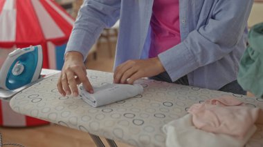Çamaşır odasındaki ütü masasında giysileri katlayan bir kadın, ev ortamını düzenli tutarken ellerini düzenli bir şekilde düzenlemeye odaklanıyor.