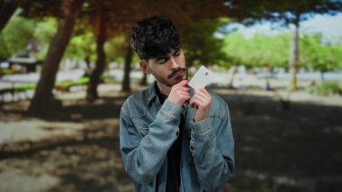 Kot ceketli genç adam güneşli bir parkta, elinde zengin yeşilliğinin ortasında düşünceli bir ifadeyle bir kredi kartı tutuyor..