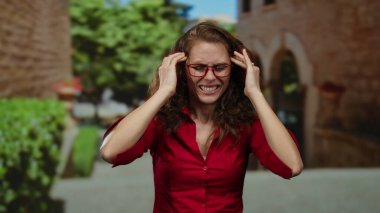 Kırmızı tişörtlü kadın şehir merkezinde hayal kırıklığını gösteriyor. Elleri başında ve gözlüklü, bulanık yeşillik ve mimariyle çevrili..