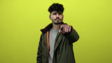 Genç adam kendine güveni olan parlak sarı bir arka plana işaret ediyor, şık yeşil bir ceket giyiyor, karizmatik ve iddialı bir tavır sergiliyor..