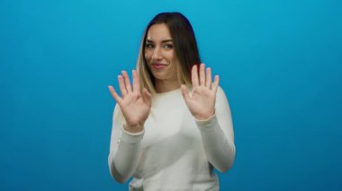 Beyaz tişörtlü genç sarışın kadın, izole bir ortamda parlak mavi bir duvara şakacı hareketlerle el hareketleriyle ve doğal bir ifadeyle duygularını ifade ediyor..