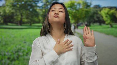 Çevresi yeşilliklerle çevrili bir parkta sadakat jesti yapan bir kadın, açık hava bağlamında genç Çinli kimliğini temsil ediyor, samimiyet ve bağlılığa odaklanıyor..