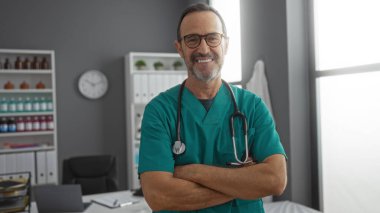 İyi organize edilmiş bir hastane odasında kollarını kavuşturup güvenle gülümseyen İspanyol erkek doktor klinik ortamda profesyonellik ve samimiyet gösteriyor..