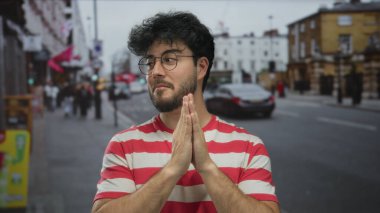 Çizgili tişörtlü, sakallı, genç İspanyol bir adam kalabalık bir şehir sokağında elleri birarada duruyor. Düşüncelerini ya da minnettarlığını doğal ışıkla ifade ediyor..