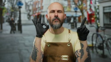 Sakallı bir adam şehir caddesinde hareketler yapıyor. Kollarında dövmeler var. Açık bir şehir ortamında moda ve yüz ifadesi sergiliyor..