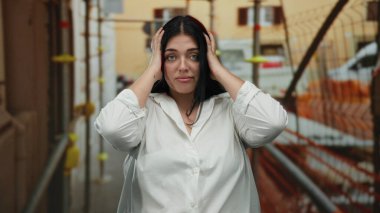 İnşaat alanındaki bir kadın stresli görünüyor, beyaz bir gömlek giyiyor, iskele ve malzemelerle çevrili, endüstriyel bir açık hava ortamında duygusal bir an yakalıyor..