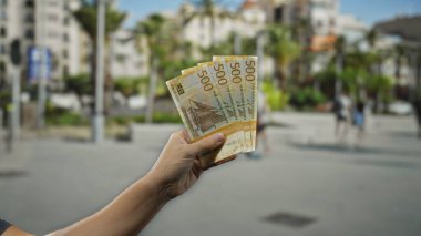Bir şehir caddesinde Norveç kronu banknotları tutan adam, canlı bir şehir zeminine karşı para sergiliyor, seyahat ve finans temalarını vurguluyor.