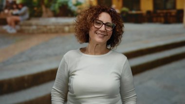 Açık şehir sokak ortamında gözlük takan, orta yaşlı gülümseyen, kıvırcık saçlı kadın, şehir yaşam tarzını ve neşeli tavrını sergiliyor, memnuniyeti ve rahatlığı vurguluyor..