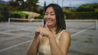 Yeşil ve manzaralı arka planla çevrili açık tenis kortunda güvenle gülümseyen genç bir kadın eğlenceli bir atmosfer olduğunu gösteriyor..