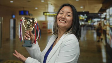 Havaalanı terminalinde gülümseyen bir ifadeyle kupayı elinde tutan kadın, kapalı bir ortamda gezginlerle ve tabelalarla başarılar sergiliyor..