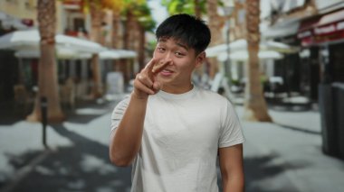 Beyaz gömlekli genç adam, güneşli bir şehir sokağında palmiye ağaçlarıyla eğlenceli bir jest yapıyor, açık hava kentsel manzarasını çekiyor..