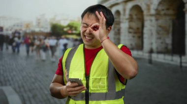 Sarı yelek giyen bir adam akıllı telefon tutarken iyi işaret veriyor. Kalabalık ve arka planda antik mimari ile dolu kalabalık bir sokak tabelası var..