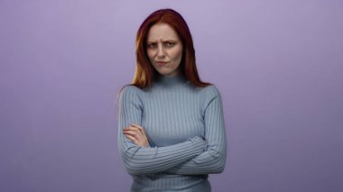 Kızıl saçlı kadın, kollarını mor arka plana doğru uzatmış düşünceli bir duruşta, hafif ışıkta durgun bir ifade sergiliyor, mavi kazak giyiyor ve sükuneti taşıyor.