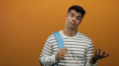 Sırt çantalı genç adam turuncu bir arka planda duruyor, düşünceli ve anlamlı görünüyor, çizgili bir gömlek giyiyor..