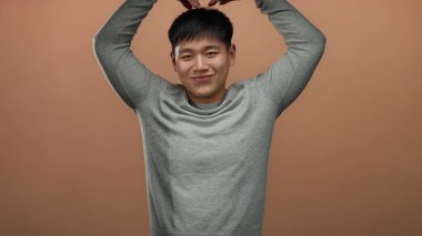 Sıradan süveter giyen genç Çinli bir adam izole edilmiş kahverengi bir arka plan duvarına şakayla poz veriyor, neşe ve pozitifliği ifade ediyor..