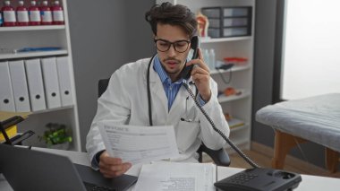 Klinikte gözlük takan genç adam telefonla konuşurken evrak işleriyle uğraşıyor. Etrafı ofis malzemeleri ve tıbbi malzemelerle çevrili. Profesyonel davranışları vurguluyor..