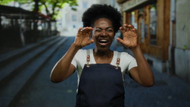 Önlük giymiş bir kadın neşeli bir şehir caddesinde poz veriyor güneşli ağaçların altında neşeli bir atmosfer yaratıyor..