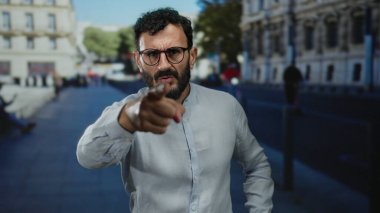 Sakallı ve gözlüklü İspanyol bir adam kalabalık bir sokakta şehir ortamında ciddi bir ifadeyle dikiliyor..