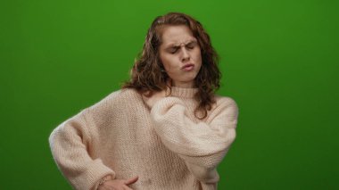 Boyun ağrısı çeken, yeşil arka planda rahat bir süveter giyen, tarafsız bir ortamda rahatsızlık veren, duruşunu ve ifadesini vurgulayan bir kadın..