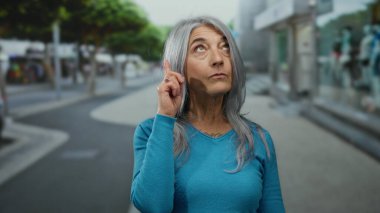 Mavi kazaklı, gri saçlı yaşlı bir kadın hareketli bir caddede düşünceli bir şekilde duruyor, açık hava kentsel ortamını merak ve güvenle işaret ediyor..