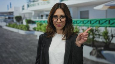 Şehir caddesinde el kol hareketi yapan bir kadın açık hava ortamını işaret ediyor. Gözlük ve ceket takıyor..