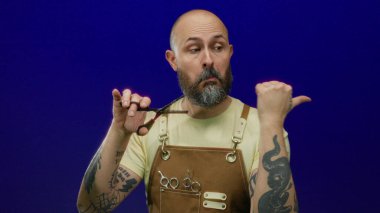 Sakallı bir erkek berber. Mavi arka planda makas tutuyor. Dövmeli, sarı tişörtlü ve deri önlüklü..