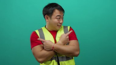 Güvenli yelek giyen genç adam, yalıtılmış yeşil arkaplanı işaret ederek yön ve güvence duygusunu gösteriyor..