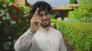 İspanyol asıllı genç bir adam parkta, parmaklarıyla şakacı bir şekilde kafasına dokunuyor, yemyeşil manzara ve canlı çiçeklerle çevrili..