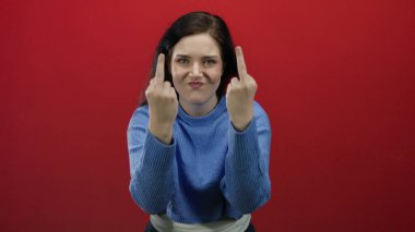 Mavi kazaklı esmer kadın canlı kırmızı arka planda duruyor. İçten duygularla çekilen hoşnutsuzluk ve meydan okuma ifadeleriyle.