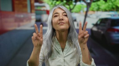 Beyaz gömlekli, kır saçlı yaşlı bir kadın sokakta dikiliyor. Ağaçların ve arabaların kentsel arka planında barış işareti var..