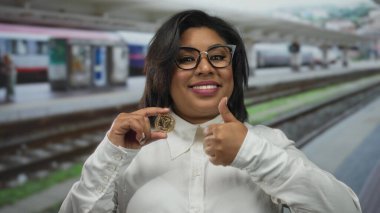 Tren istasyonundaki genç Latin kadın elinde kripto para tutuyor ve açık hava ortamında şifreli bir başarı sergiliyor..