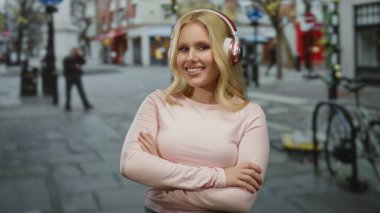 Şehir merkezinde güvenli bir şekilde duran, kulaklık takan, günlük pembe tişörtlü, neşeli bir kadın, şehir manzarası arka planında şehir ortamının tadını çıkarıyor..