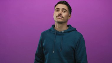 Deniz mavisi kapüşonlu genç İspanyol adam canlı mor arka plana karşı duruyor, izole bir ortamda günlük tarz ve güven yayıyor..