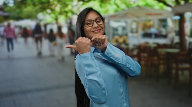 Mavi gömlekli genç bir kadın güneşli bir restoran sahnesinde canlı bir terasta bulanık insanlarla gülümserken başparmaklarını geriye doğru gösteriyor.