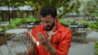 Hapishane tulumlu İspanyol adam, arka planda boş masalar olan bir lokantada kelepçeli bir şekilde perişan görünüyor..