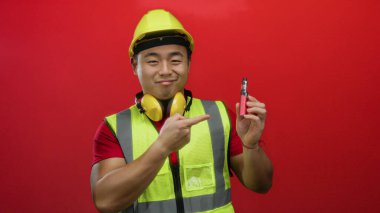 Güvenlik teçhizatlı genç adam kırmızı arka plana karşı vape cihazını işaret ederek güvenli bir şekilde neşeli bir ifadeyle ürünü gösteriyor.