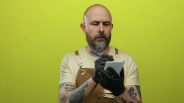 Dövmeli kel bir adam parlak sarı bir arka plan üzerine küçük bir deftere notlar yazar. Siyah eldivenler ve bej bir gömlek giyer..