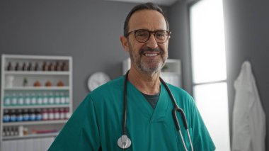 Hastane odasında gülümseyen, steteskoplu önlük giyen, klinik ortamlarda güven ve deneyim sergileyen olgun İspanyol bir adam.