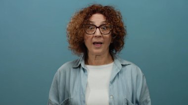 Orta yaşlı İspanyol kadın, kısa kıvırcık esmer saçlı, gözlüklü ve günlük kıyafetli, izole edilmiş mavi arka plana şaşırdığını ifade ediyor..