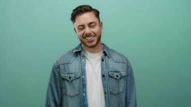 Sakallı, kot gömlekli genç adam, izole edilmiş yeşil arka plana gülümsüyor. Sıradan bir tarz ve pozitif enerji yansıtıyor.