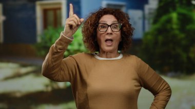 Şehir manzarası ile çevrili düşünceli bir yüz ifadesiyle İspanyol kadın, canlı bir açık hava ortamında gözlük ve günlük giysiler giyiyor.