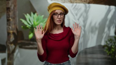 Gözlüklü ve sarı şapkalı bir kadın arka planda bitkilerle dikiliyor. Rahat ve kendine güvenen kırmızı elbiseli bir kadın..