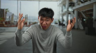 Şehir sokaklarında duygusal olarak el kol hareketi yapan genç Çinli bir adam, gri bir kazak giyiyor, arka planda modern şehir mimarisi var..