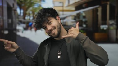 Sakallı genç bir adam, kalabalık bir caddede, sakin bir şehir ortamında güvenle gülümserken bir telefon konuşması yapıyor..