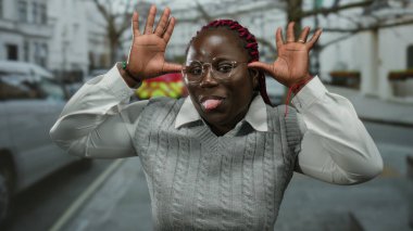 Şehir caddesinde komik suratlar yapan bir kadın, gözlüklü ve gri kazaklı, arka planda ağaçlar ve arabalar olan, açık havada neşeli ve neşeli ifadeler sergiliyor..