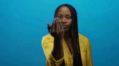 Mavi bir duvarın önündeki odak noktasını işaret eden kadın, sarı bir gömlek giyiyor, özgüven ve yoğunluk yayıyor, jest ve ifadeyi vurguluyor..