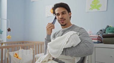 Yakışıklı bir adam yatak odasında bebek kucağında telefonda konuşuyor. Evin içinde beşik ve dekorasyonla sıcak bir aile ortamı yaratıyor.
