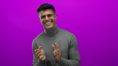 Genç adam neşe ve karizma saçan canlı pembe arka plana karşı gözlük ve kazak içinde mutlu bir şekilde alkışlıyor.